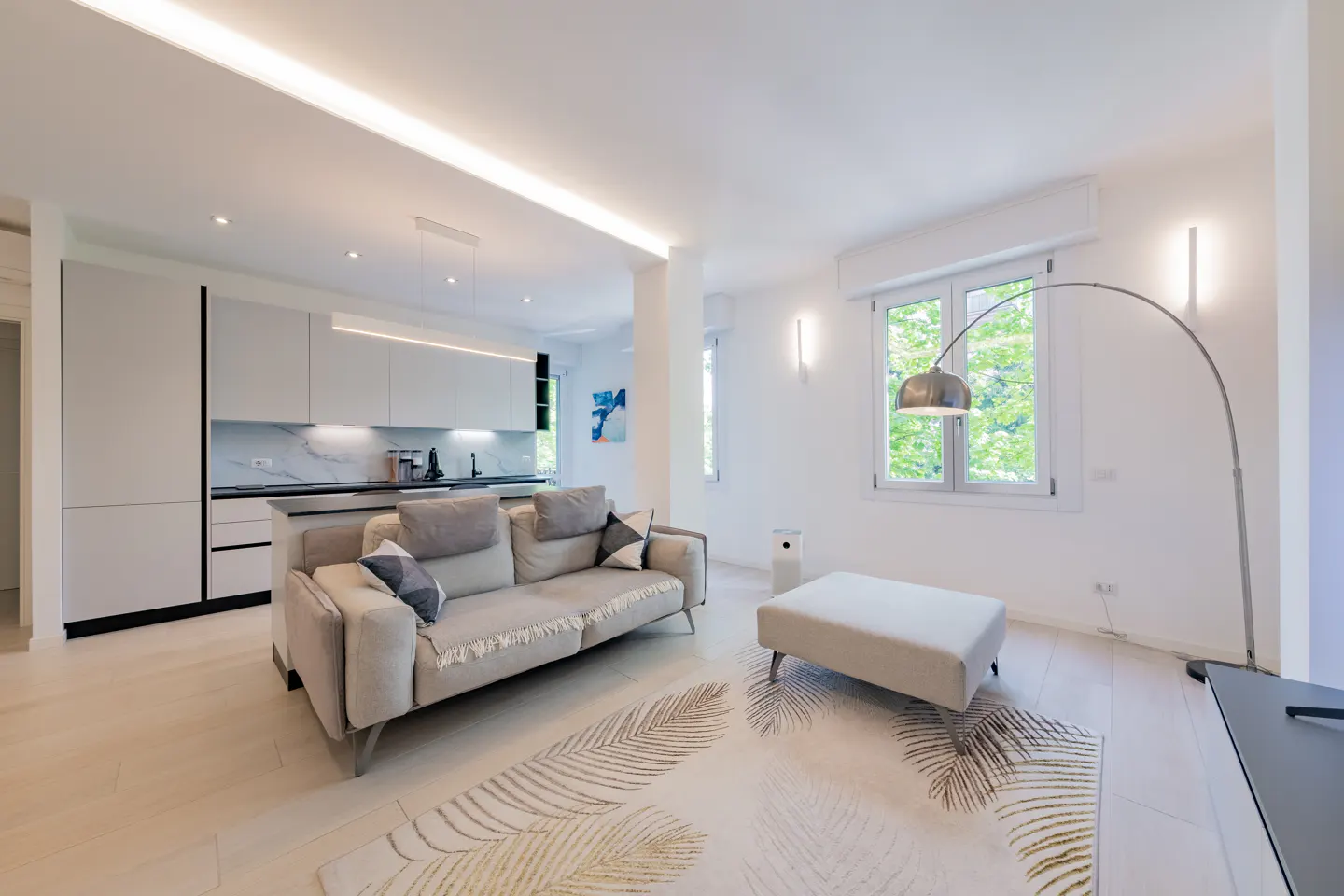 Bright, modern living room with a gray sofa, ottoman, and leaf-patterned rug. White kitchen in the background, natural light from the window.