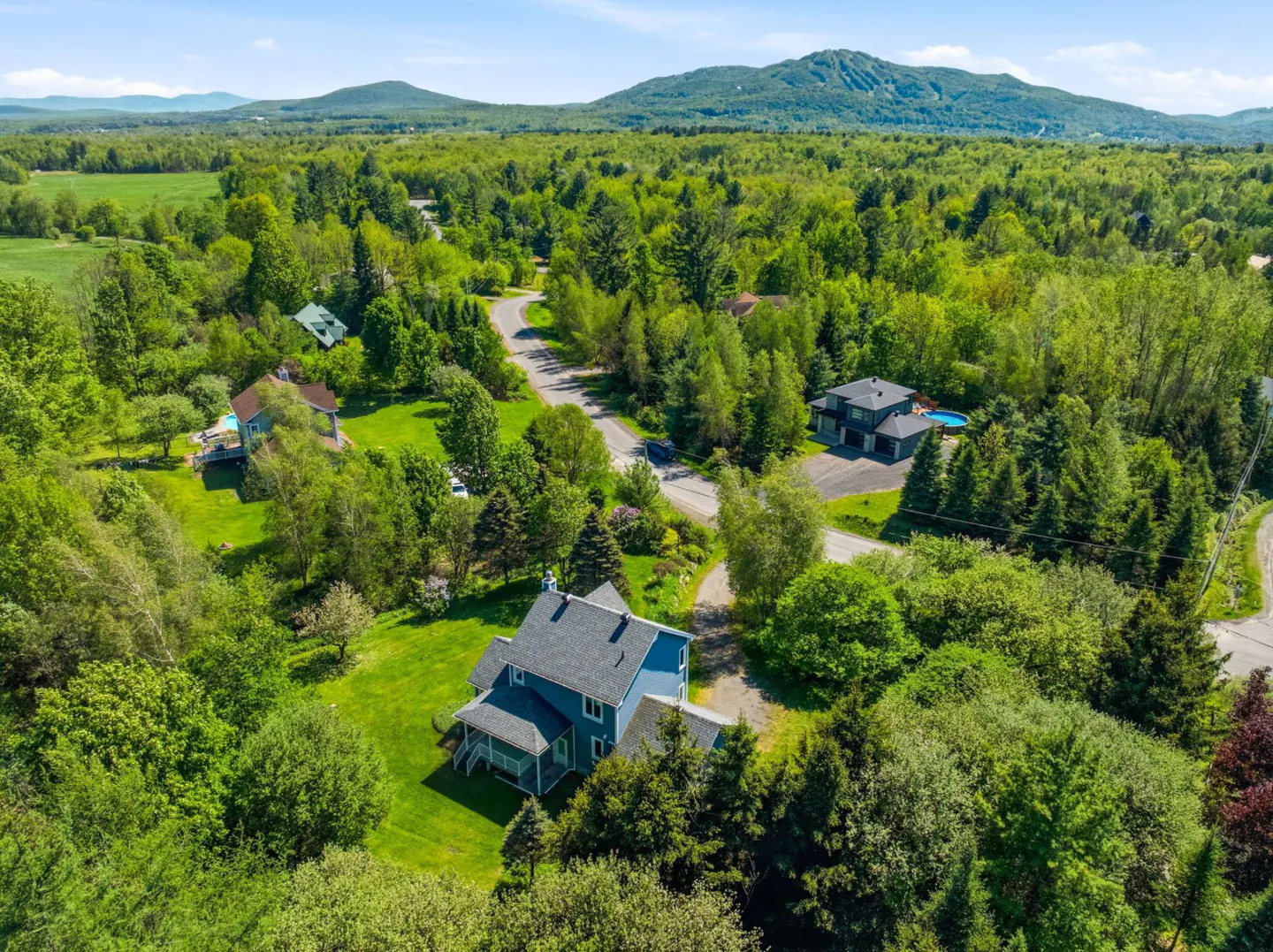Aerial view of a blue house nestled in a lush, green forest landscape with mountains in the distance under a blue sky.