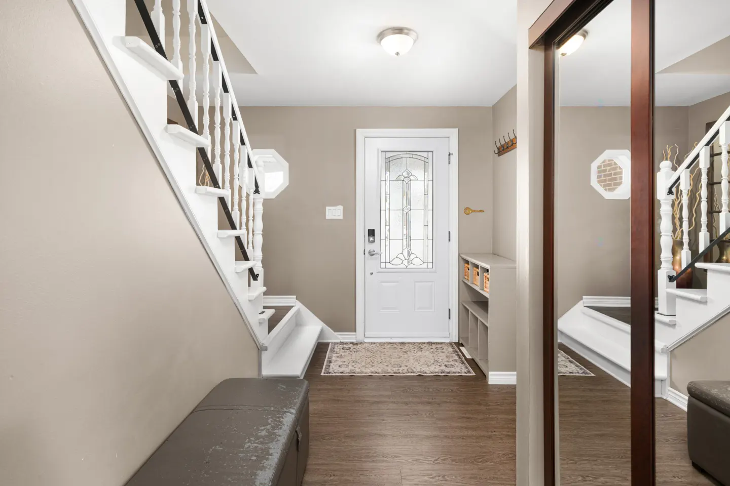 Foyer with white door, staircase, and brown wood floors. A gray bench sits near the stairs, and a mirror reflects the scene.