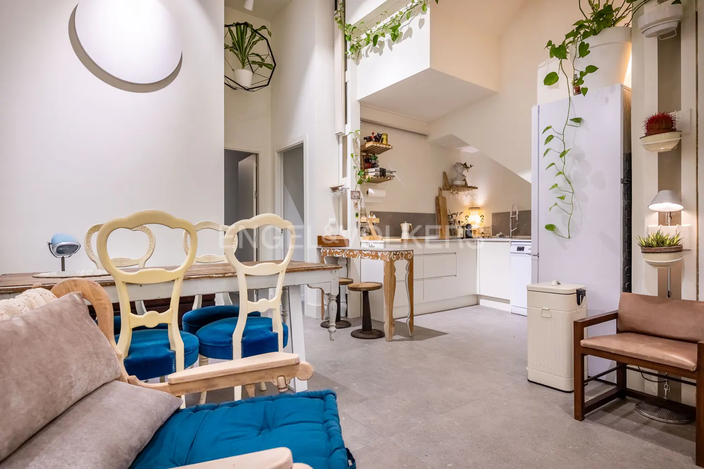 Bright, modern apartment interior with white walls, wooden table and chairs with blue cushions, and a white kitchen in the background. Green plants add a natural touch.