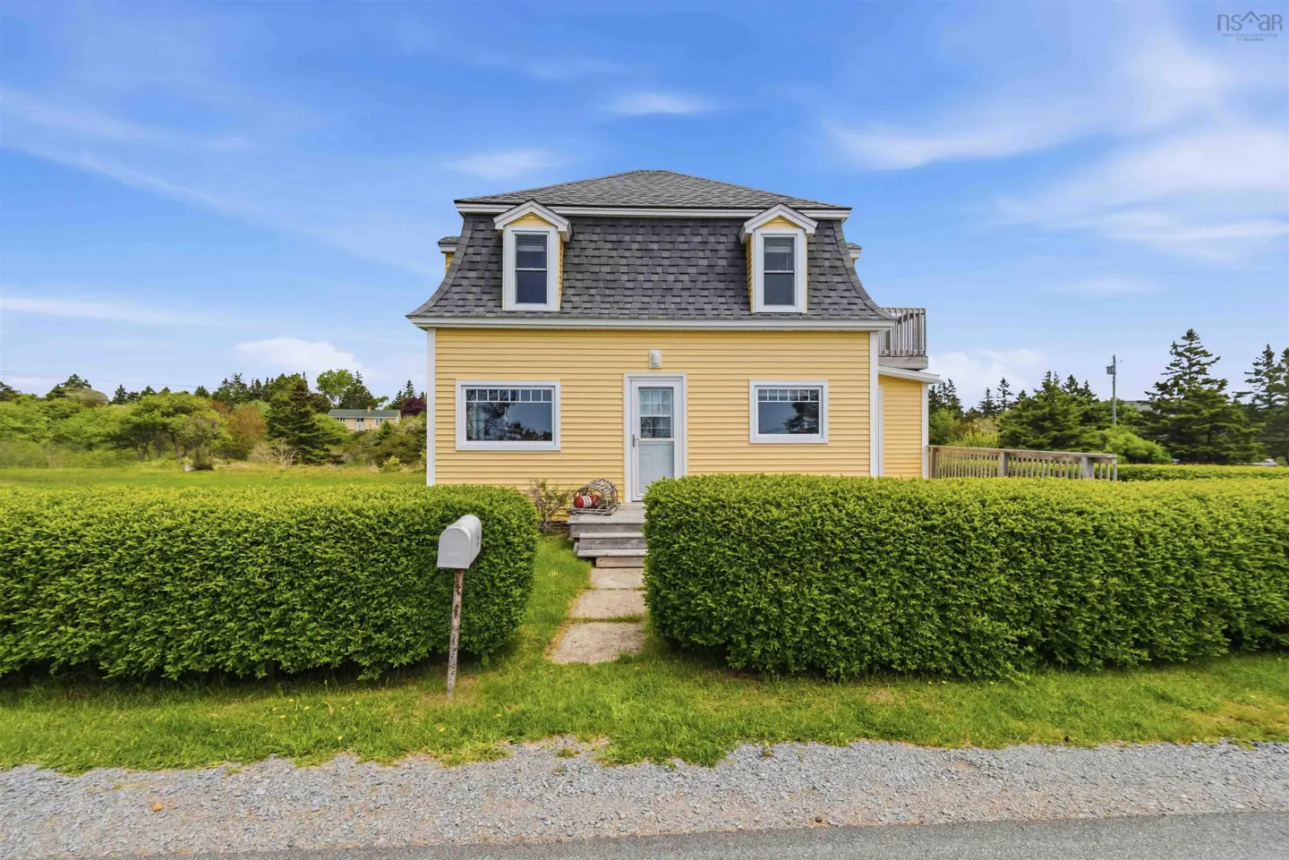 A yellow two-story house with a grey shingled roof, surrounded by tall green hedges and a blue sky.
