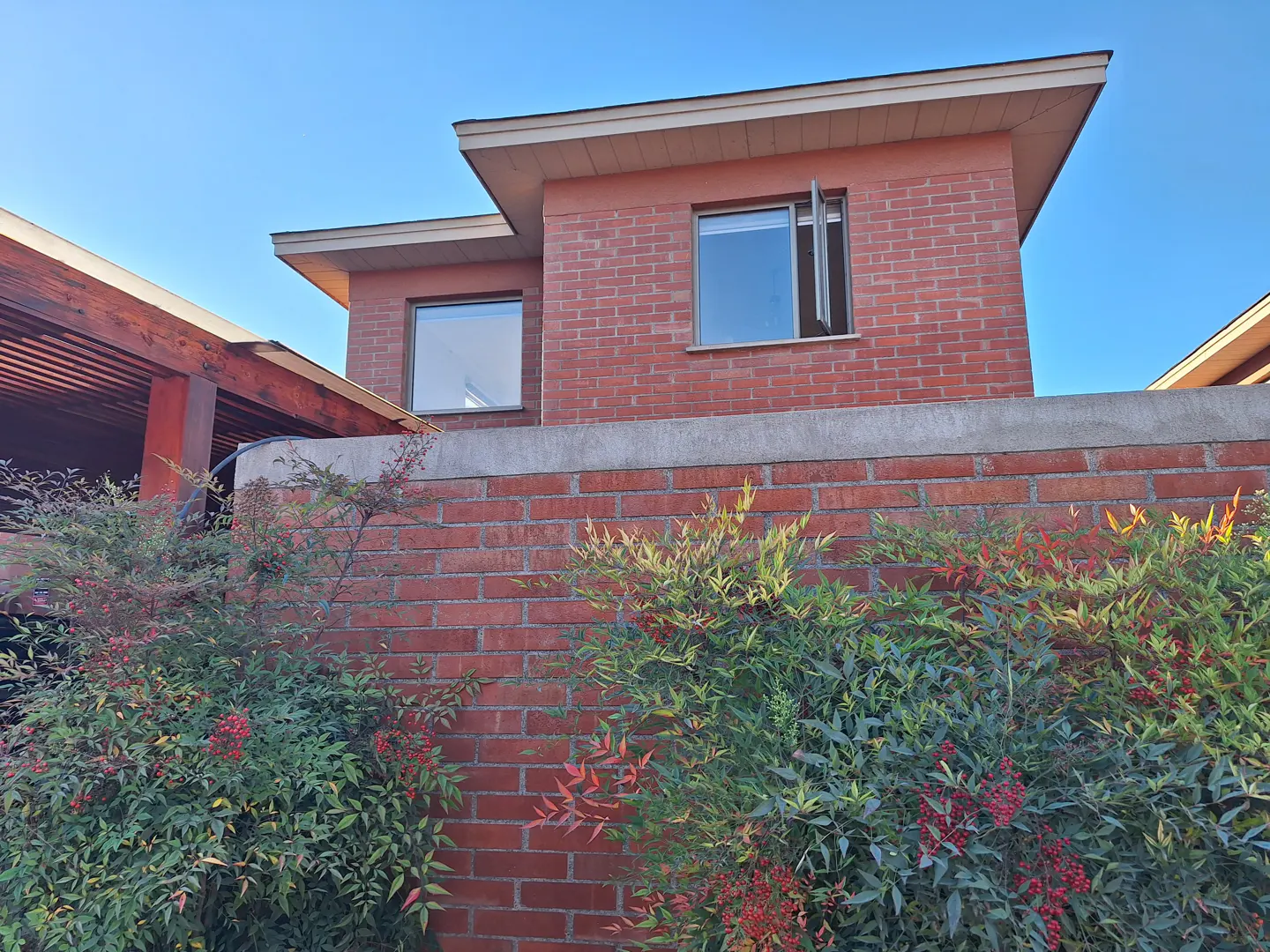 Exterior view of a red brick house with two windows, one slightly open, and a brick wall with green bushes in front.