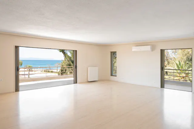 A bright, empty room with beige walls and marble floors. Open sliding doors reveal a balcony with a view of the ocean and beach.