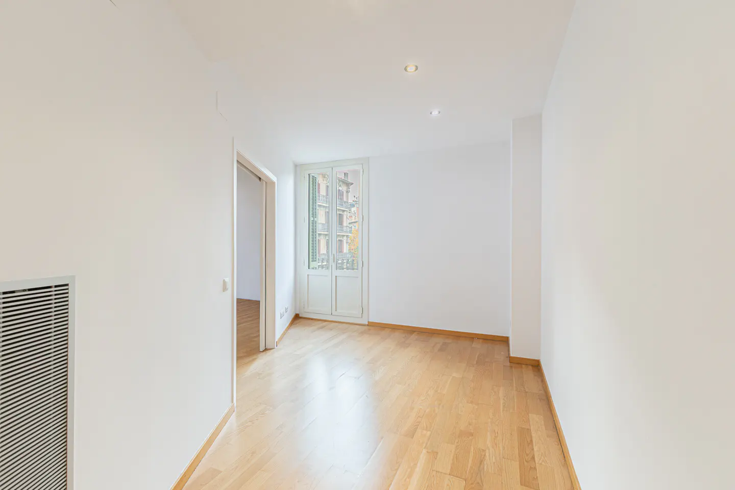 Empty room with white walls, light wood flooring, and a glass door leading to a balcony overlooking a city street.