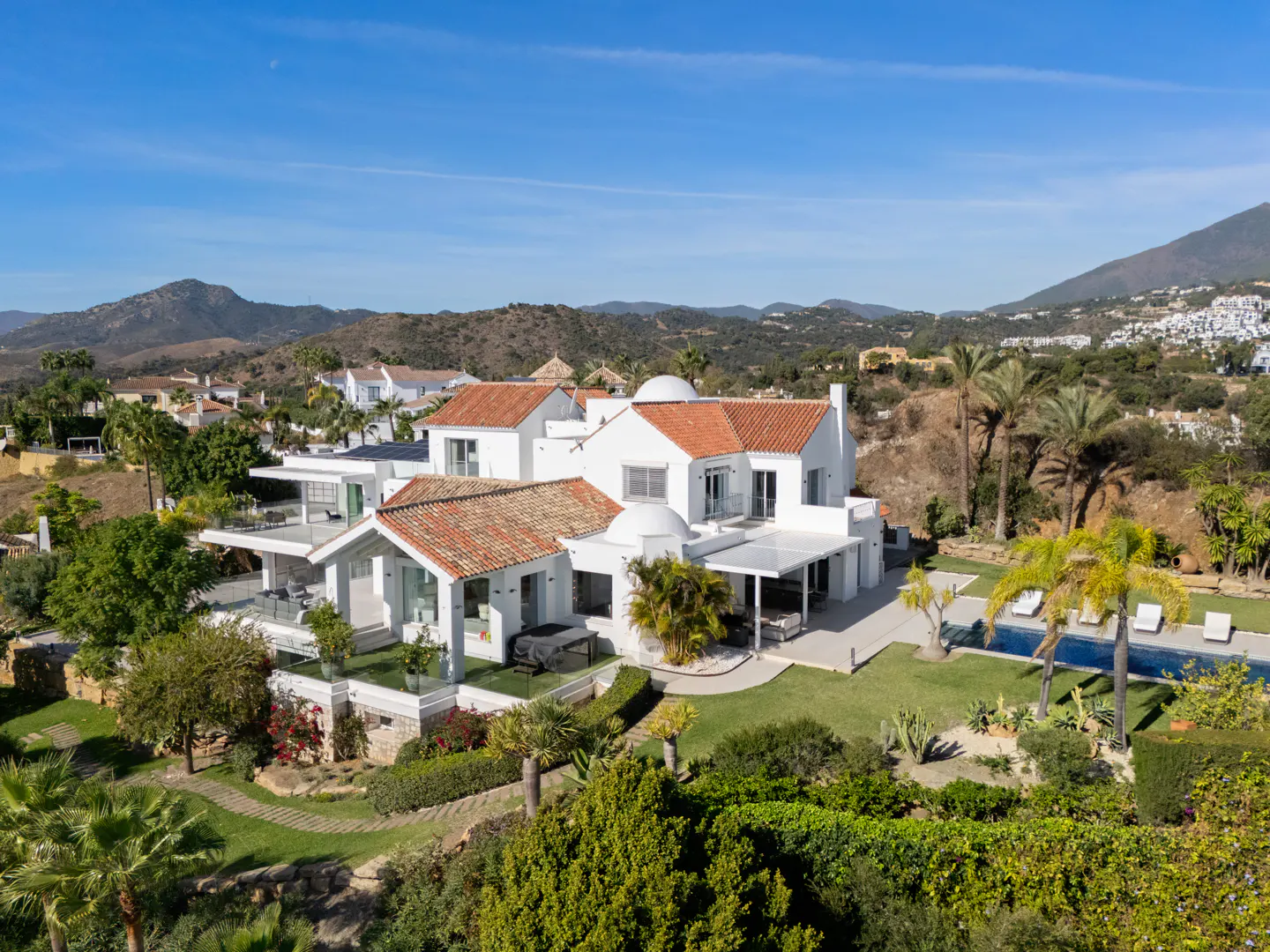 Aerial view of a white villa with a red tile roof, a pool, and lush green landscaping against a blue sky.