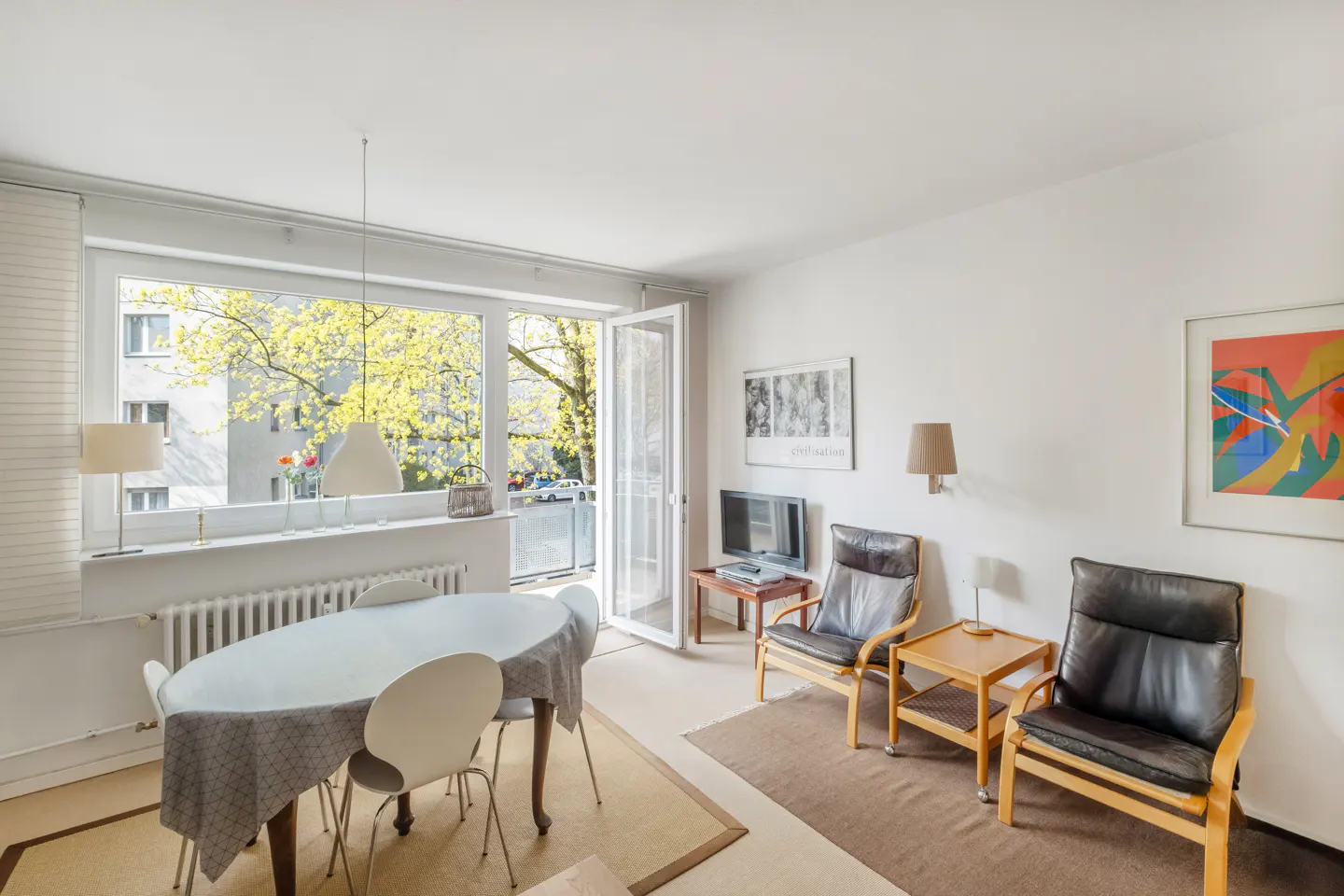 Bright living room with dining table, chairs, and two leather armchairs. Balcony door open to a view of trees. Artwork on the white walls.