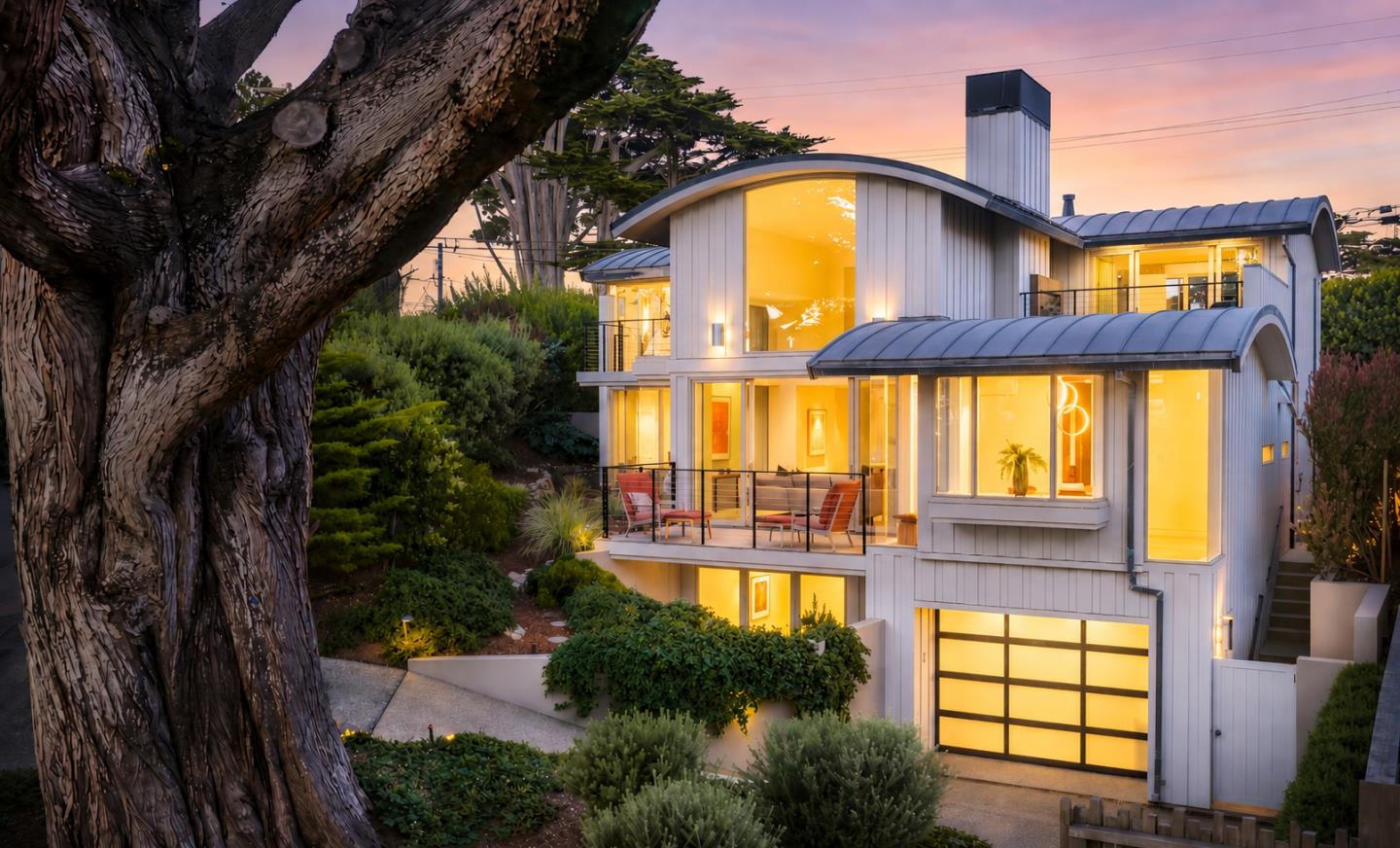 Modern multi-story home with curved roof, glowing interior lights, and a balcony at sunset, framed by a large foreground tree.