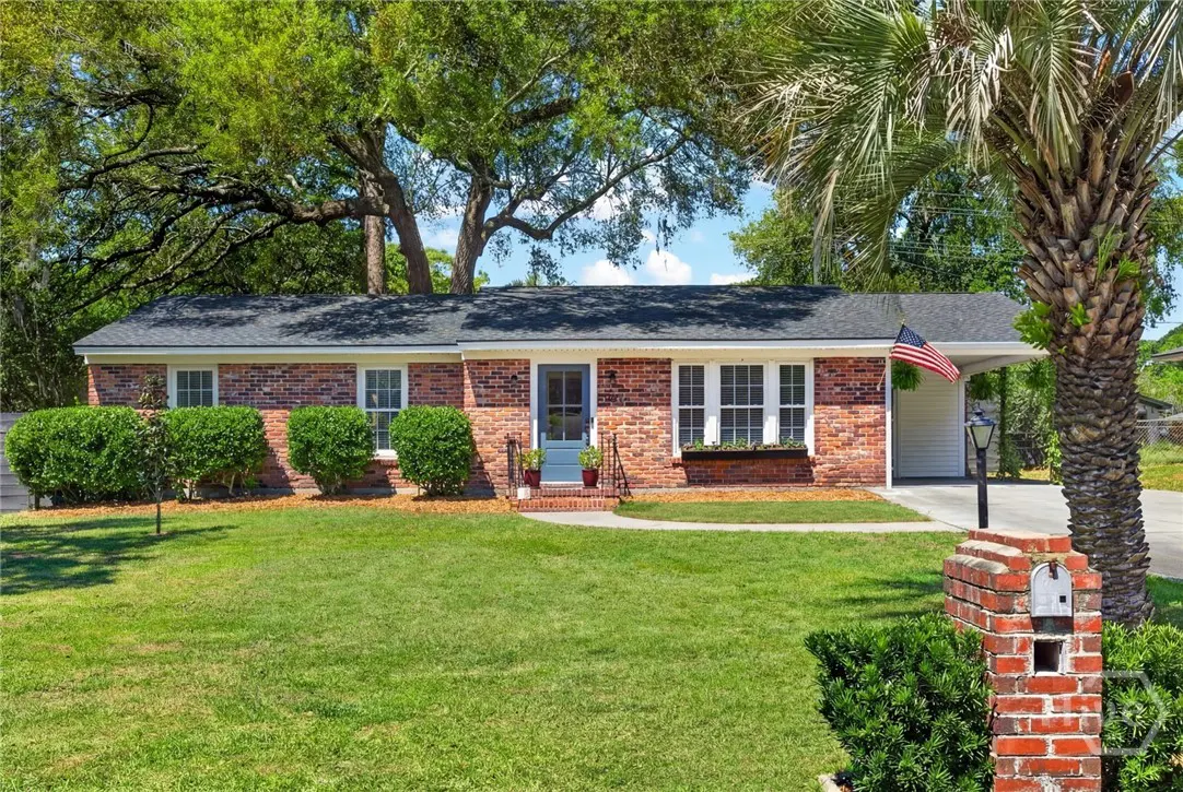 A red brick house with a dark roof, green lawn, and a palm tree on the right. An American flag hangs near the carport.