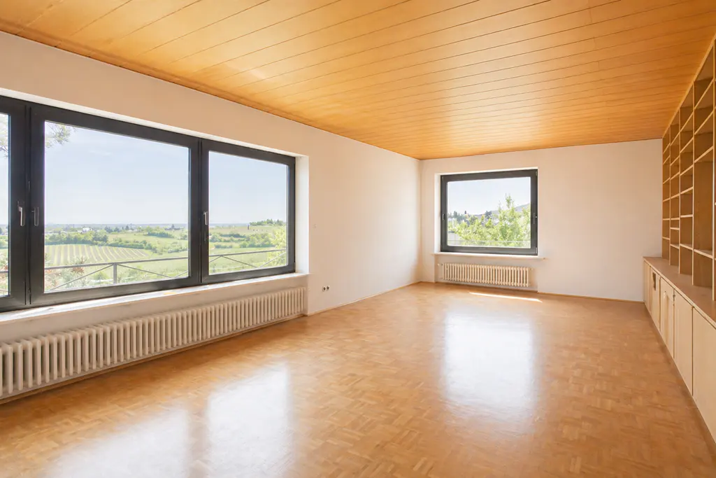 Empty room with wood floors, white walls, and a wood plank ceiling. Large windows with black frames overlook a green landscape. Built-in shelves on the right wall.