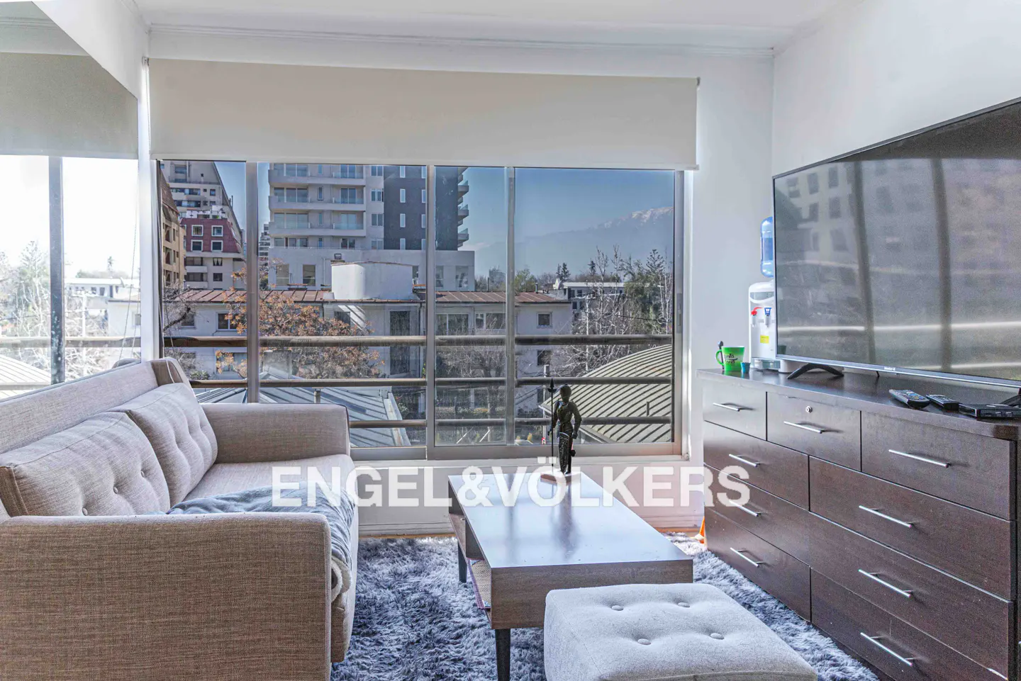 Living room with a gray sofa, coffee table, and ottoman on a gray rug. Large windows show city views. A TV sits on a dark wood dresser.