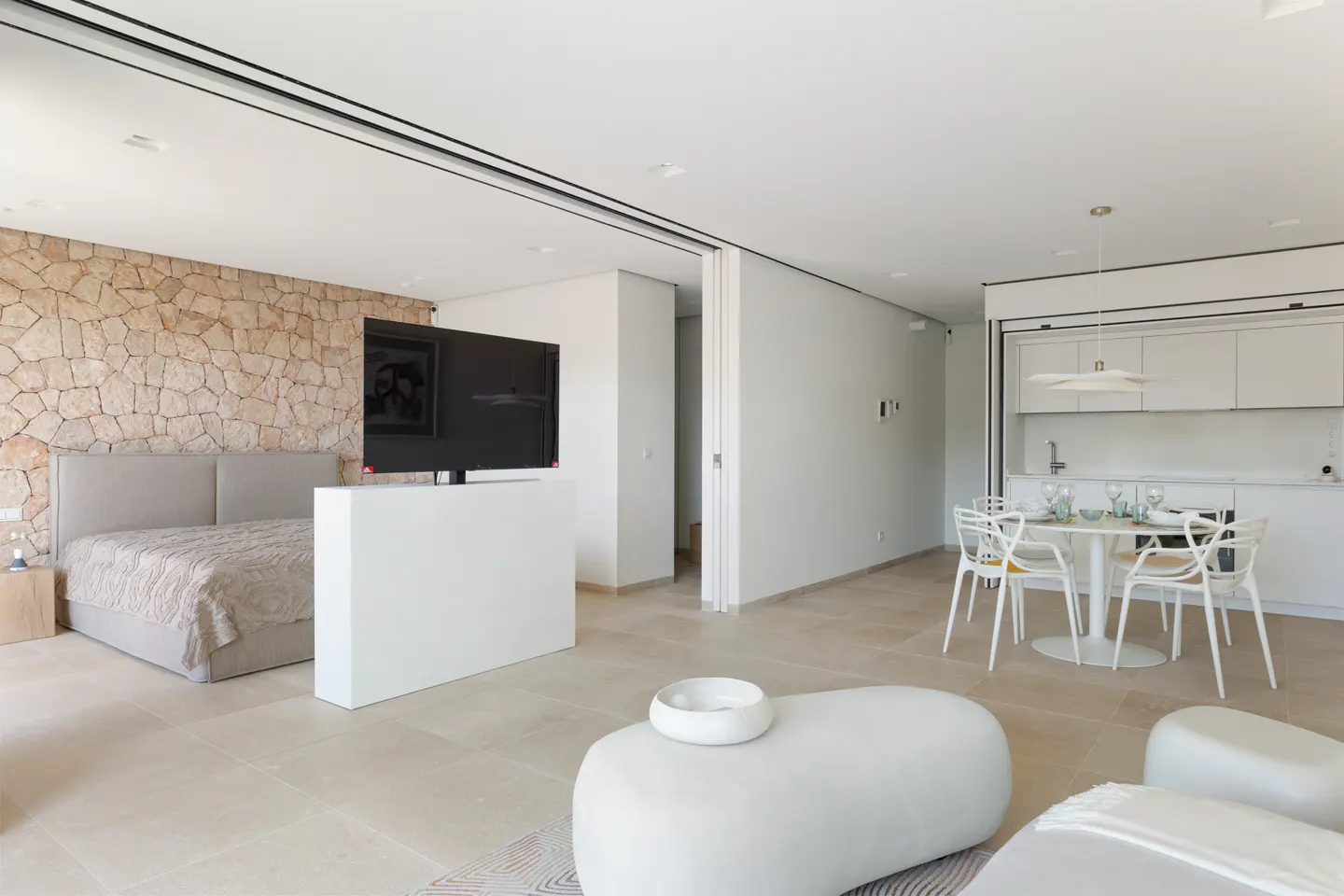 Open-concept living space with a bedroom, TV, dining area, and kitchen. The bedroom has a stone wall. The color palette is mostly white.