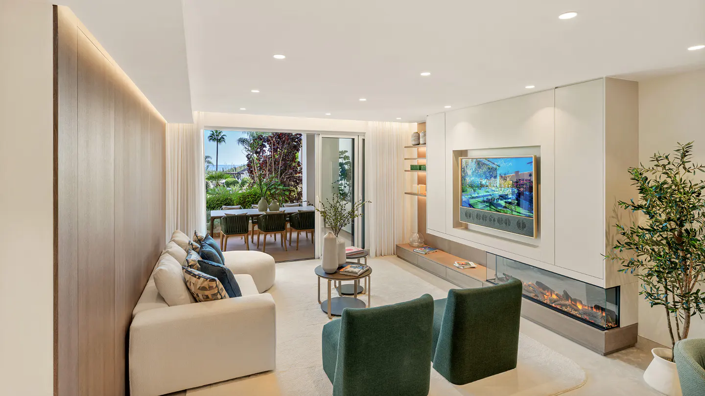 Bright living room with white sofa, green chairs, and a TV above a modern fireplace. Outside, a patio table and lush greenery are visible.