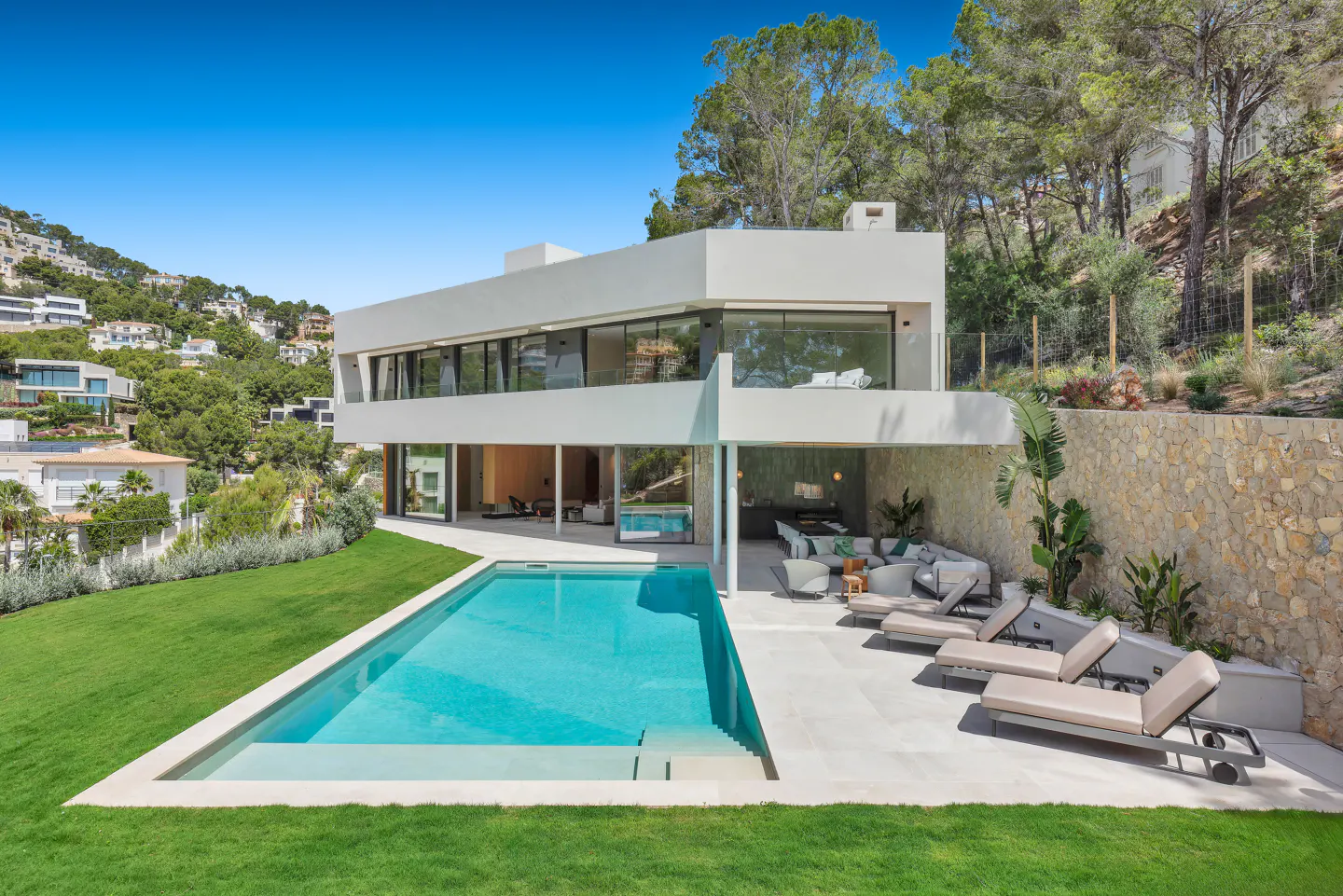 Modern white house with a pool, lawn, and lounge chairs. The house has large windows and is surrounded by trees.