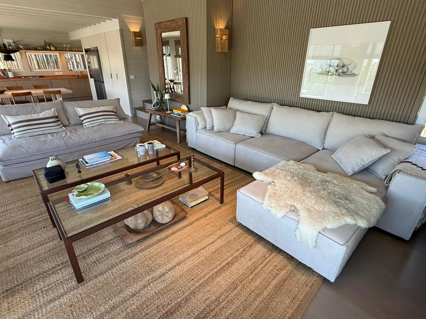 Living room with beige sofas, a jute rug, and a glass-topped coffee table. A large mirror and artwork adorn the textured wall.