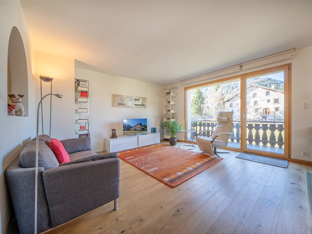 Bright living room with grey sofa, orange rug, recliner chair, and large glass doors opening to a mountain view.