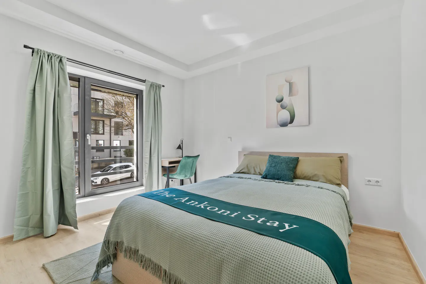 A bright bedroom with a bed, desk, and window. The bed has a light green blanket and a dark green runner with "The Ankoni Stay" written on it.
