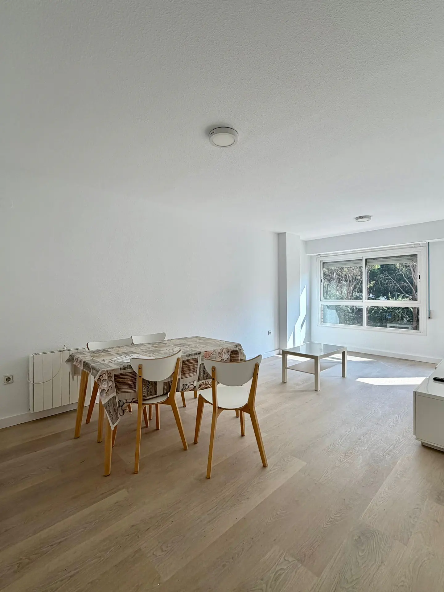 Bright, empty room with wood floors, white walls, and a large window. A table with chairs sits near a radiator, and a coffee table is near the window.
