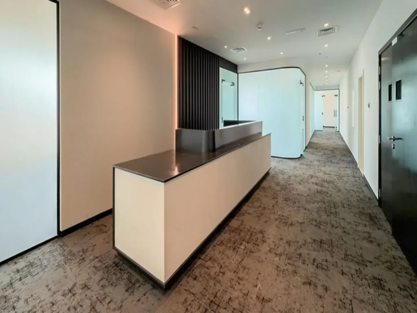 A modern office lobby with a white reception desk, black accents, and a long hallway with doors.