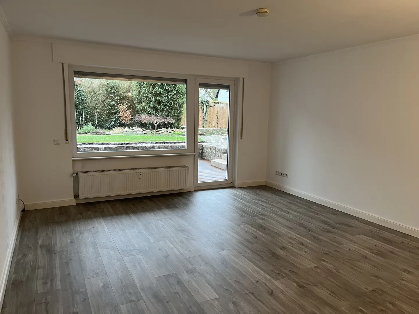 Empty room with gray wood floors, white walls, and a large window overlooking a green garden with a stone wall and patio.