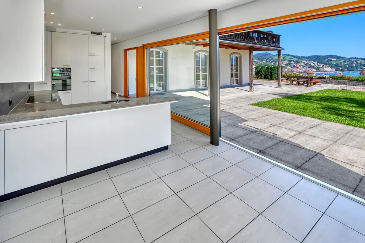 A modern kitchen with white cabinets and a gray countertop opens to a patio with a view of a lake and town.