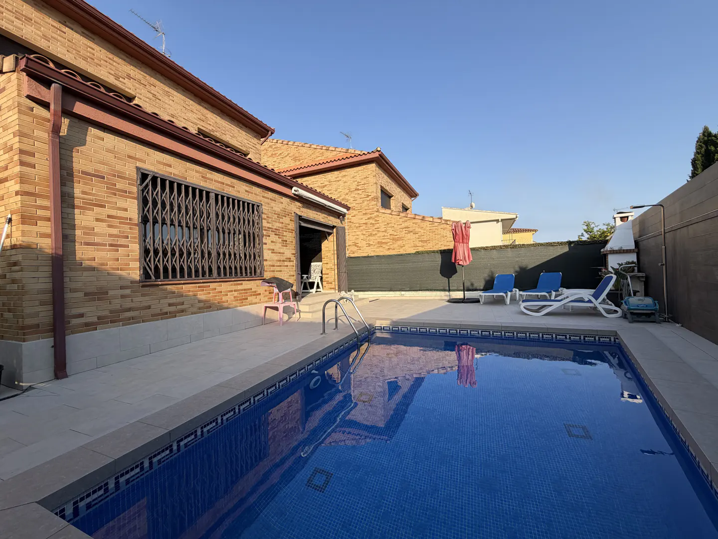 Backyard view of a brick house with a blue tiled pool, lounge chairs, and a pink umbrella under a clear blue sky.