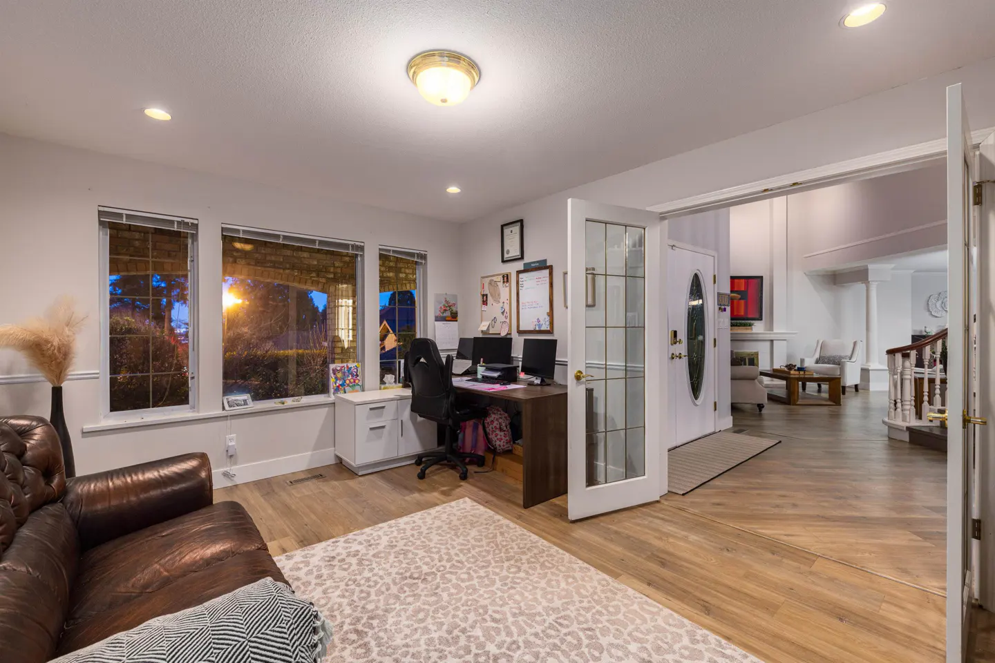 Bright, open-concept home office with a brown leather couch, desk, and large windows. A doorway leads to a living room with a staircase.