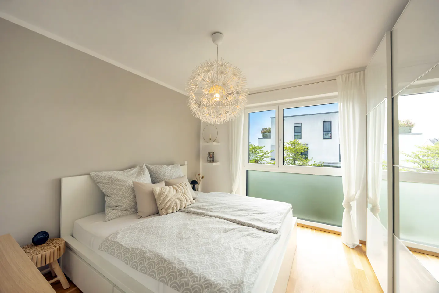 Bright bedroom with a white bed, patterned bedding, and a dandelion-shaped light fixture. A large window offers a view of a modern building.