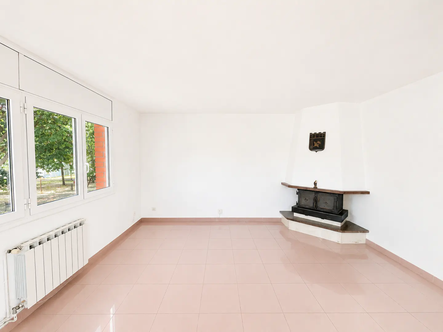 Empty room with white walls, pink tiled floor, large windows, a white radiator, and a corner fireplace with a black insert.