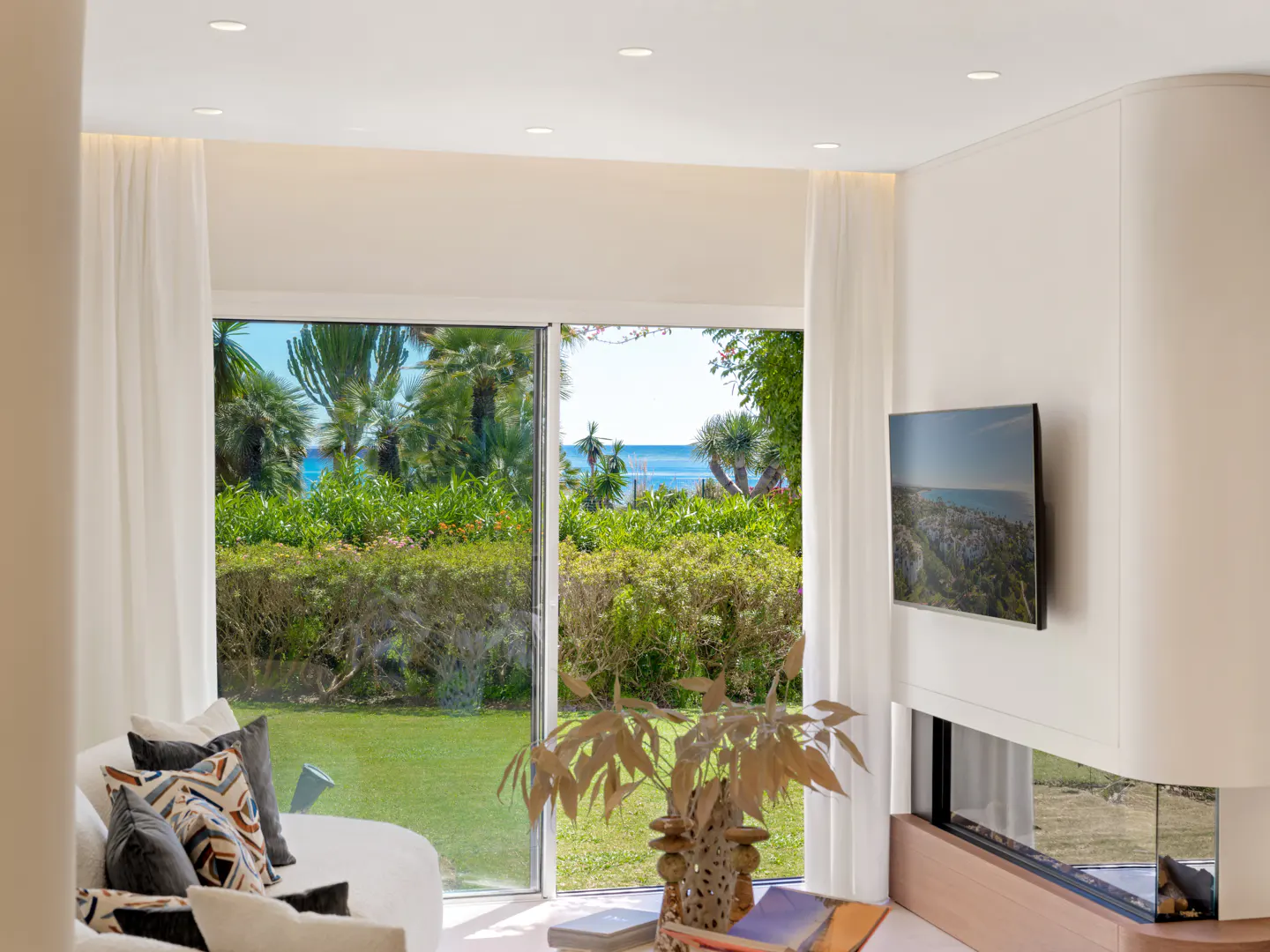Bright living room with white sofa, pillows, and a view of a green lawn, trees, and the ocean through a large window. A TV hangs above a modern fireplace.