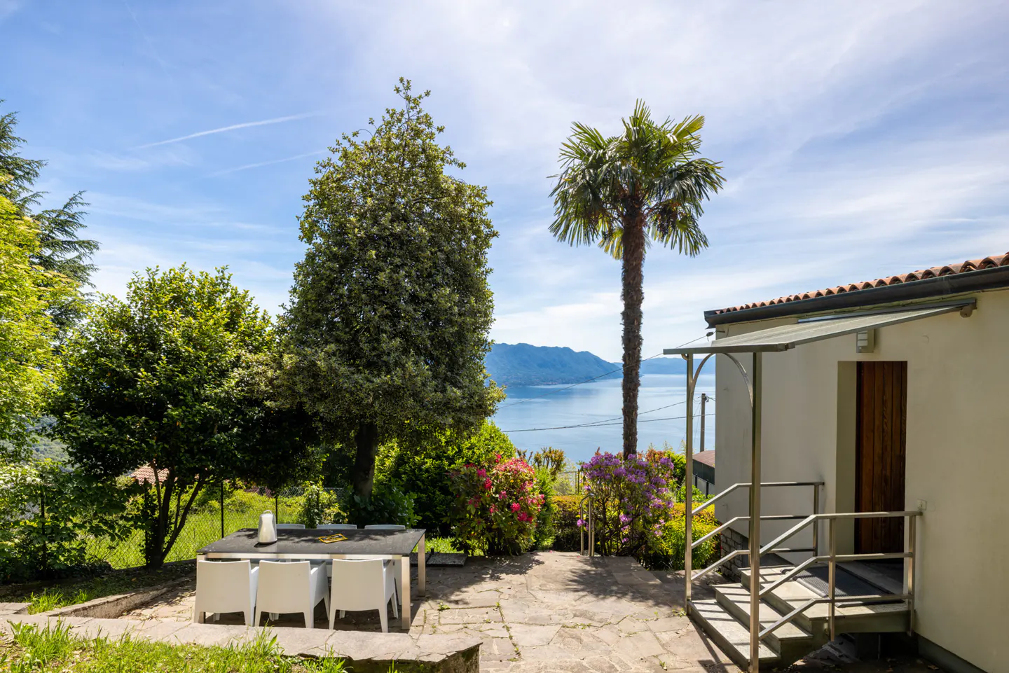 Outdoor patio with table, chairs, and view of lake. Palm tree and lush greenery surround the stone patio. Blue sky with light clouds.
