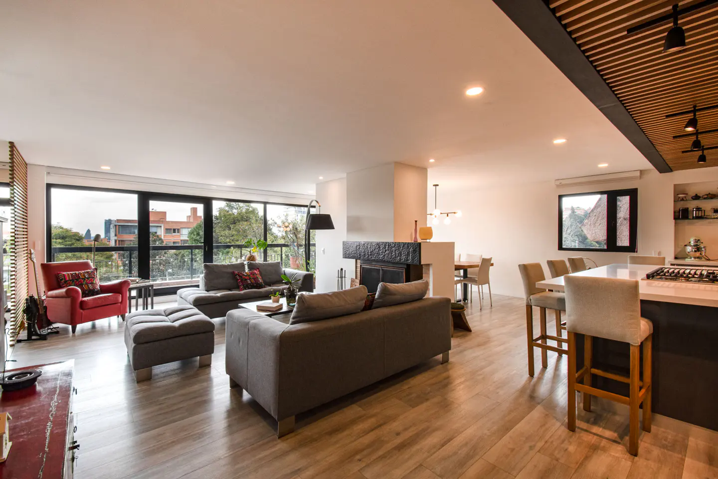Open-concept living space with gray sofas, red armchair, fireplace, and kitchen island with bar stools. Large windows offer city views.