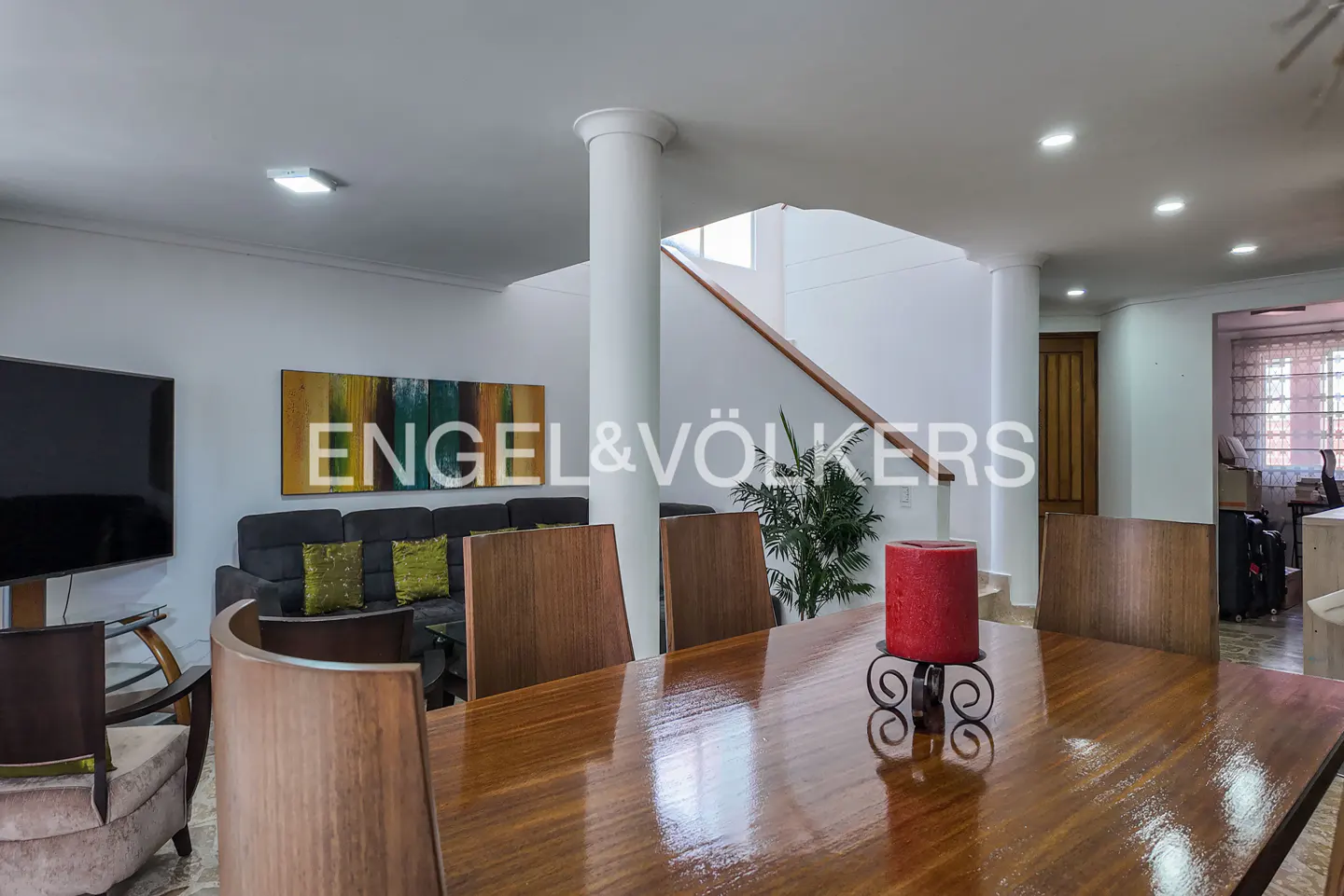 Dining room with a polished wood table, red candle centerpiece, and chairs, leading to a living area with a sofa and stairs.