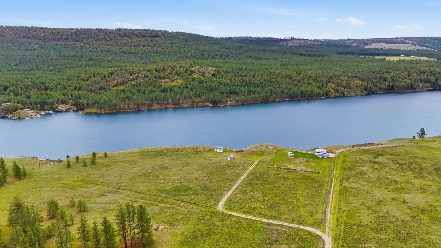 Aerial view of a green field with a blue river, forest, and a few small buildings under a blue sky.