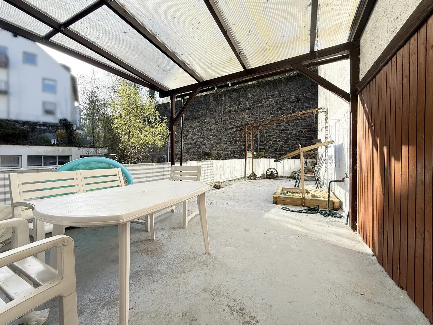 Outdoor patio with a white table and chairs under a translucent roof. A bench, a sandbox, and a stone wall are in the background.