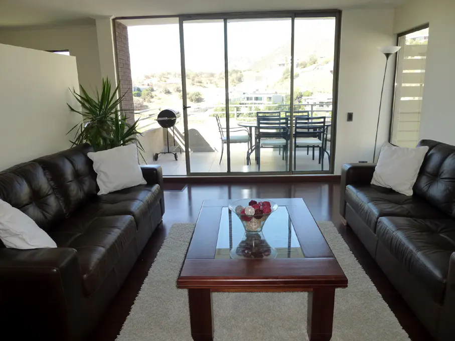 Living room with two brown leather sofas, a glass-topped coffee table, and a balcony with a grill and outdoor seating.