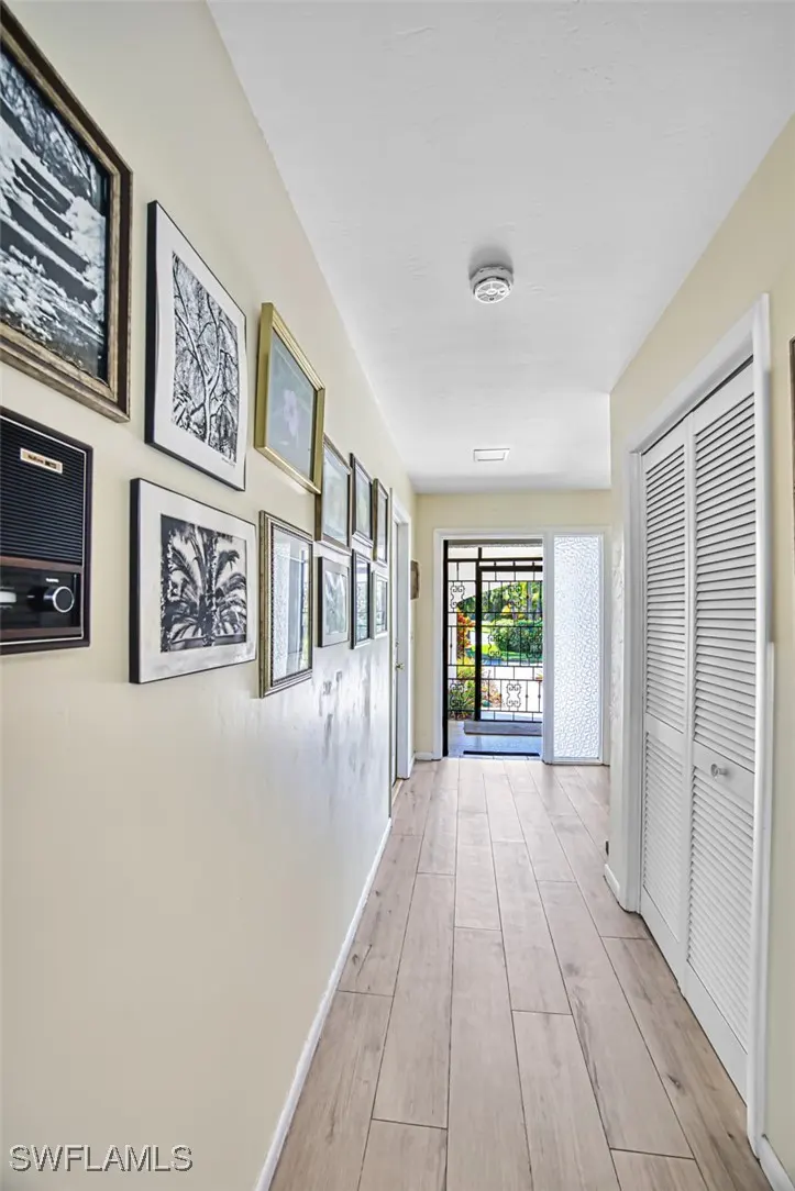 Hallway with light wood-look tile, cream walls, and white trim. A gallery wall of framed art lines the left. Double louvered doors are on the right. View to the outside.
