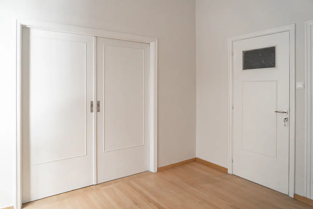 Bright room with light wood floors, white walls, and two white doors. One is a sliding double door, the other has a glass panel.