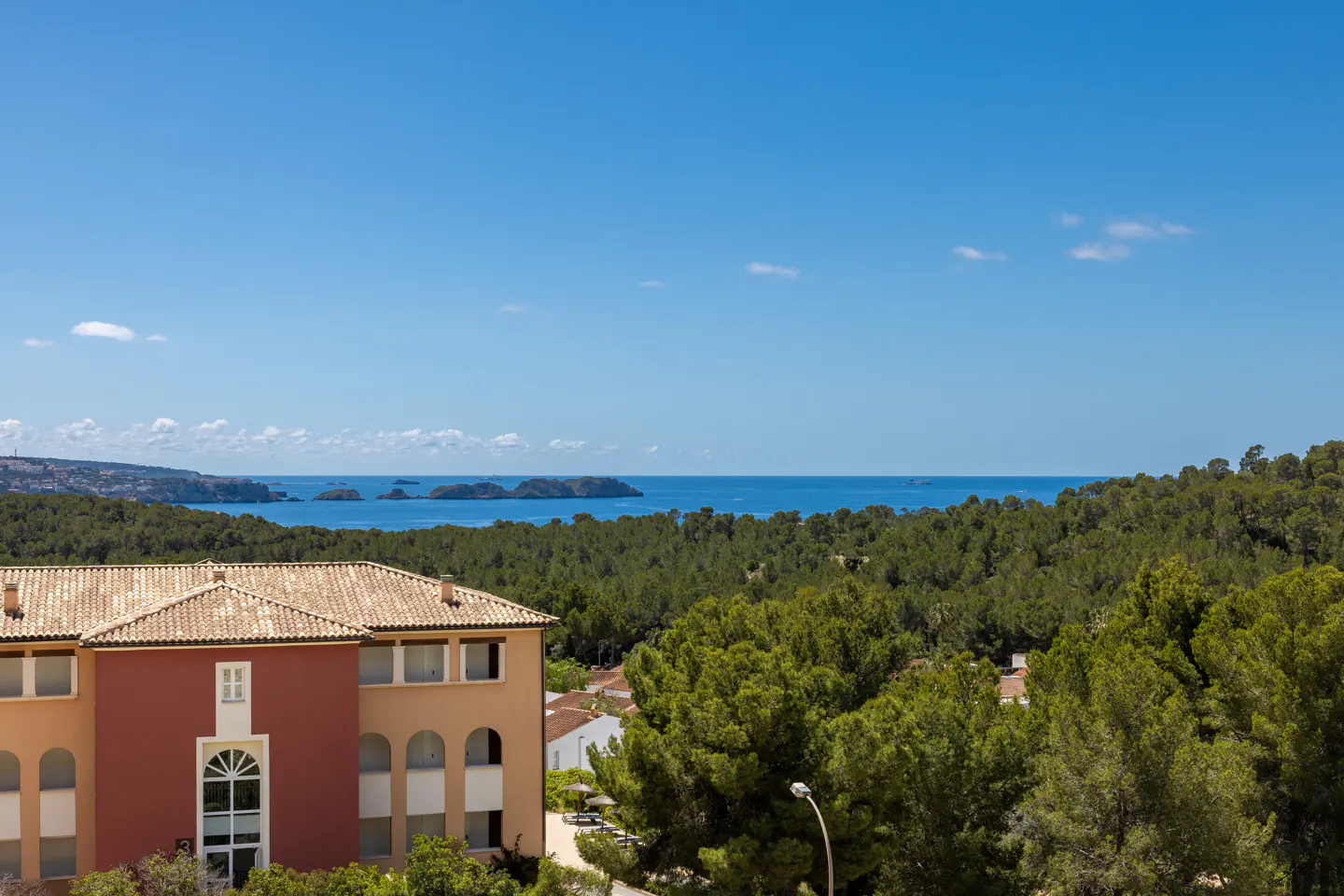 View of a building with a tan roof, green trees, and the ocean under a blue sky.