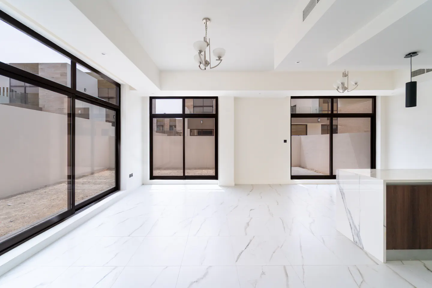 Bright, empty room with white marble floors, white walls, and black-framed windows. Chandeliers hang from the ceiling.
