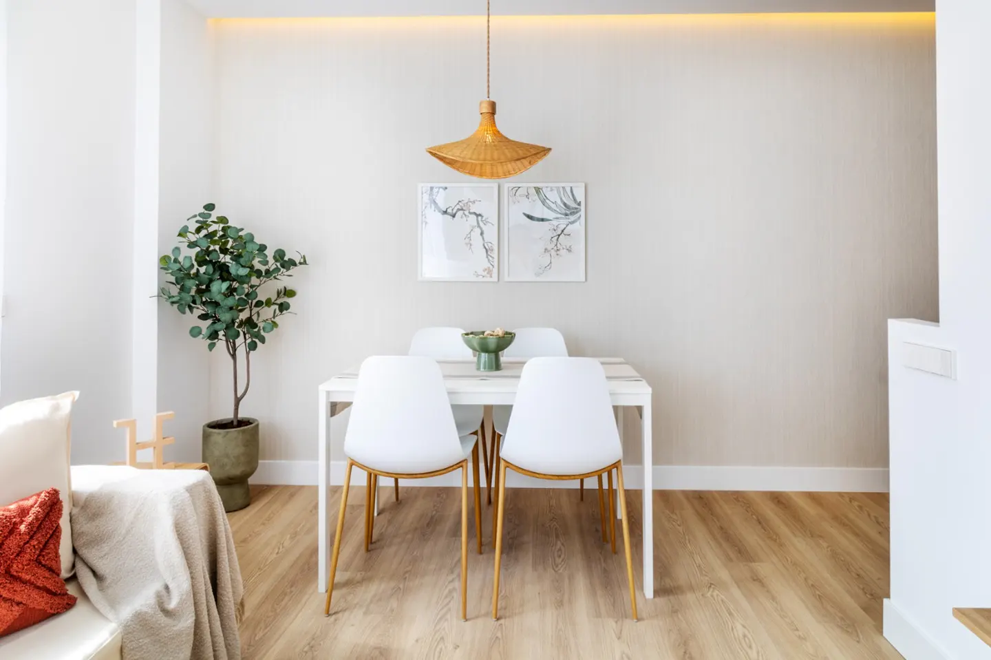 Bright dining area with a white table, four white chairs with gold legs, and a woven pendant light. Two framed prints hang above.