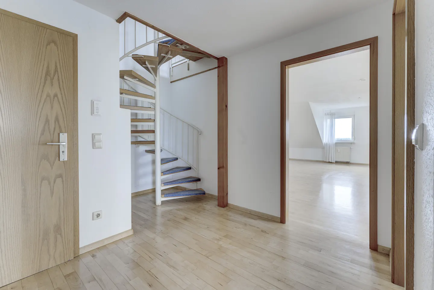 Hallway with light wood floors, white walls, and a spiral staircase. A wood door is on the left, and an open doorway leads to another room.