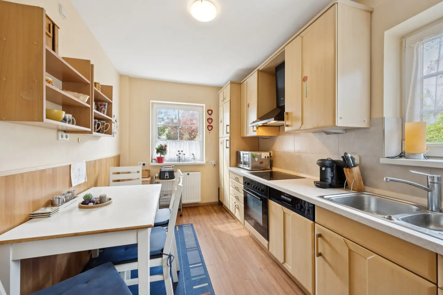Bright kitchen with light wood cabinets, white countertops, and a dining table with blue cushions. A window overlooks a garden.