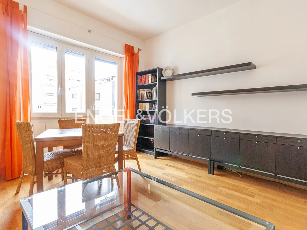 Living room with wood floors, dining table with wicker chairs, black shelving unit, and orange curtains.