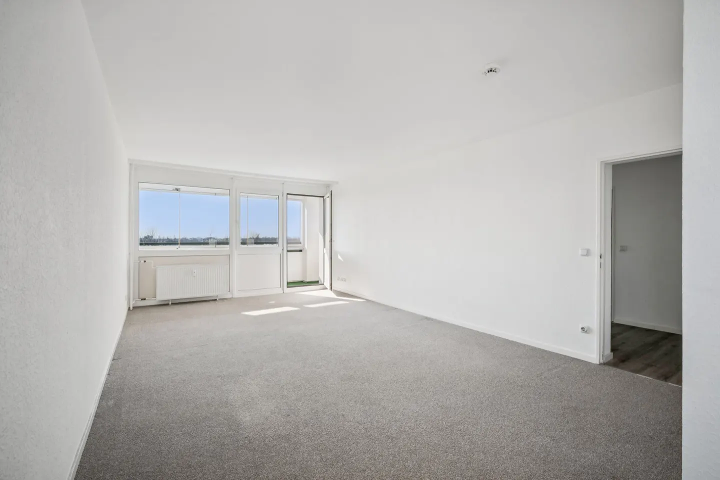 Bright, empty room with gray carpet, white walls, and a balcony with a view. A doorway leads to another room.