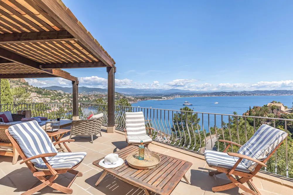 Outdoor patio with wooden furniture, striped cushions, and a pergola overlooking a blue ocean and distant mountains on a sunny day.