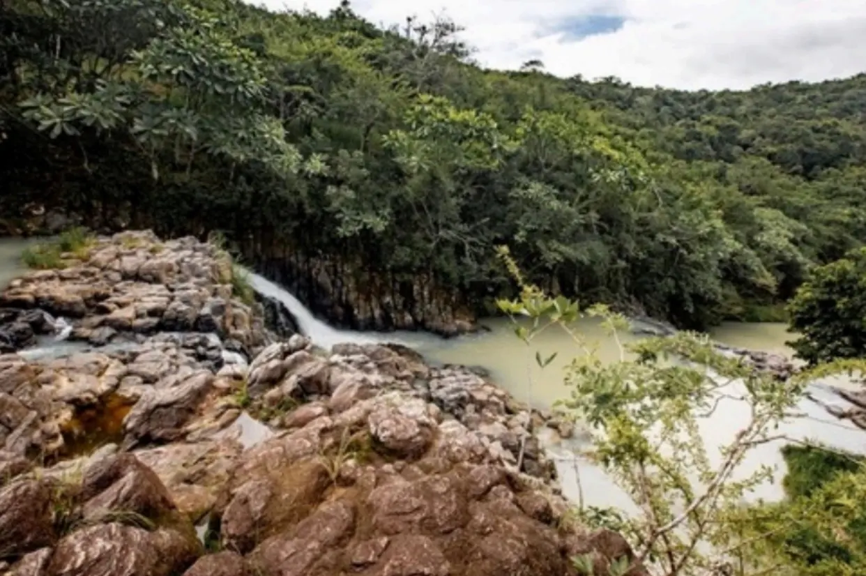 A scenic view of a small waterfall cascading over rocks into a river, surrounded by lush green trees.