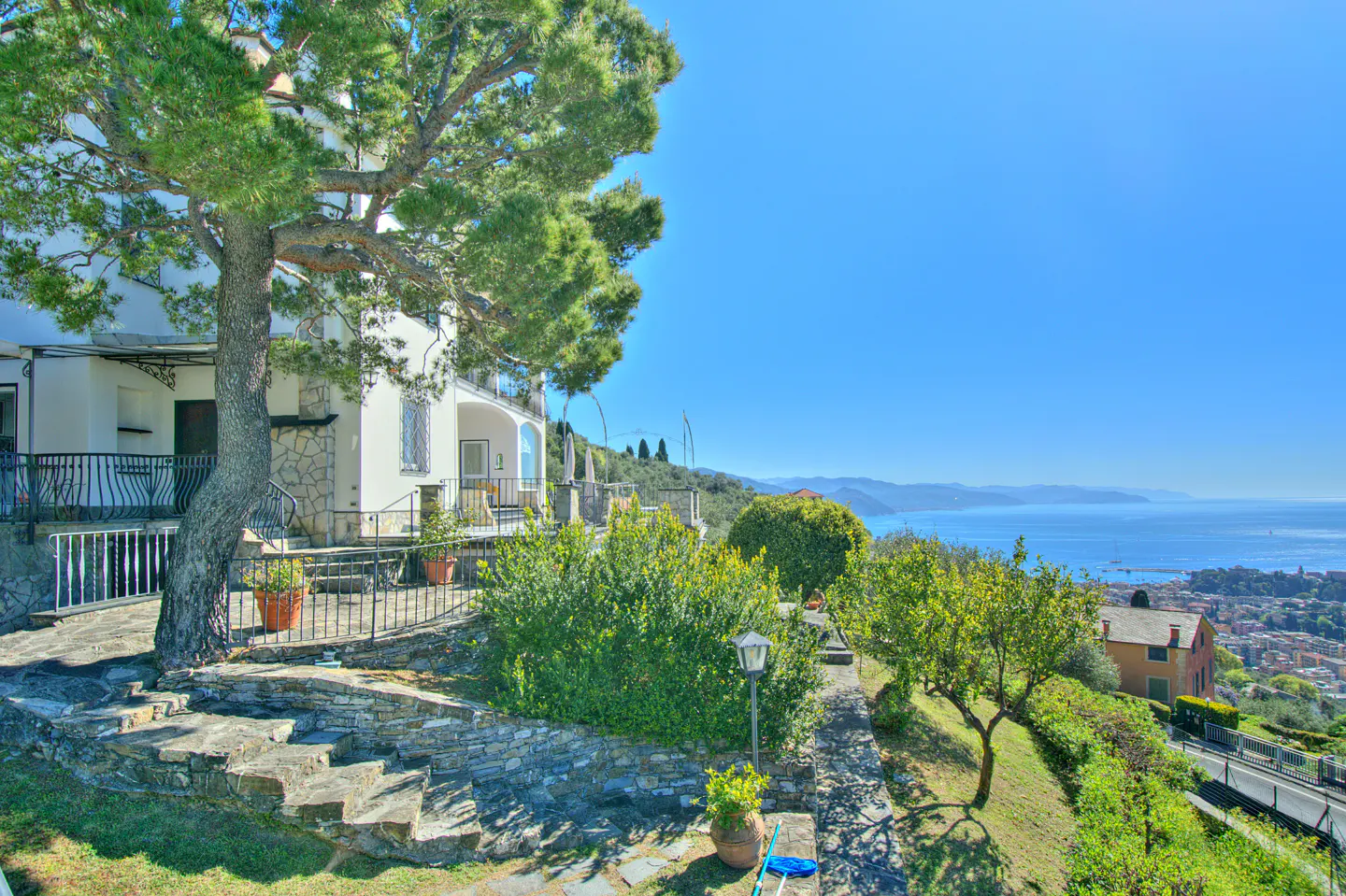 Exterior view of a white house with a balcony, stone steps, and a large tree overlooking the ocean under a clear blue sky.