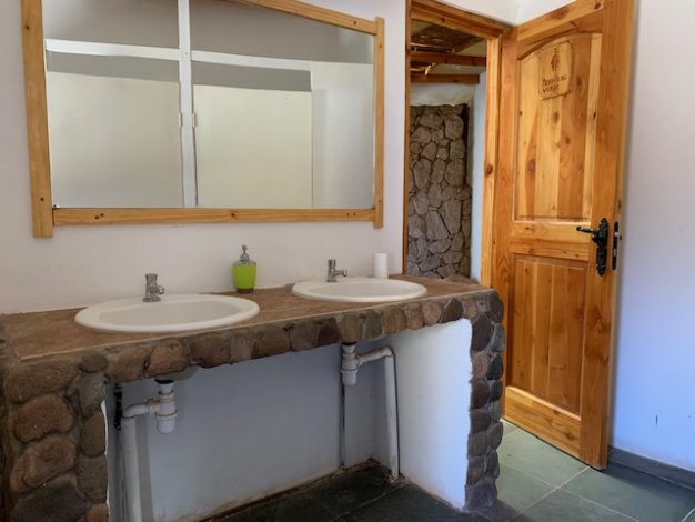 Rustic bathroom with double sinks, stone counter, wood-framed mirror, and open wooden door to a stone-walled stall.