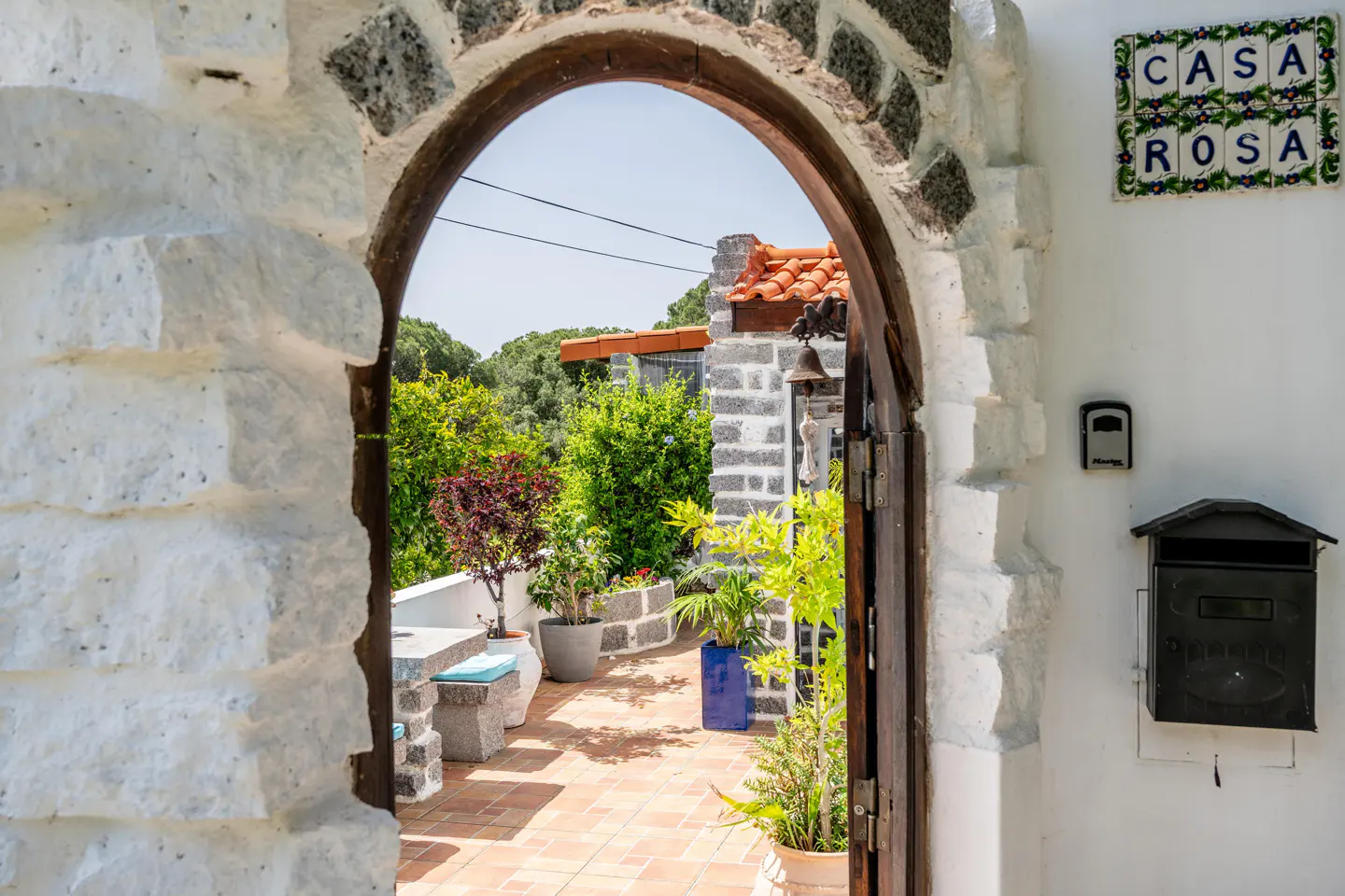 View through stone archway to a patio with plants, a stone bench, and a view of trees. "Casa Rosa" tile, mailbox, and doorbell on white wall.