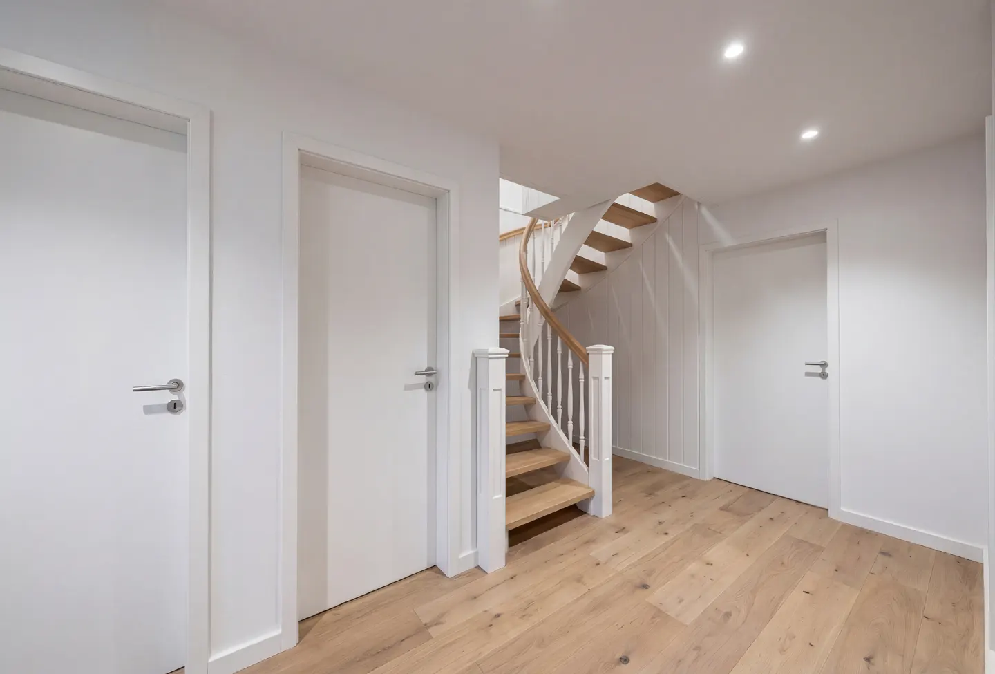 Bright hallway with wood floors, white walls, and three white doors. A wooden spiral staircase with white railings is visible.